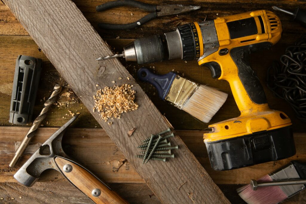 DIY home improvement tools including a drill, paintbrush, screws, and wood plank on a workbench